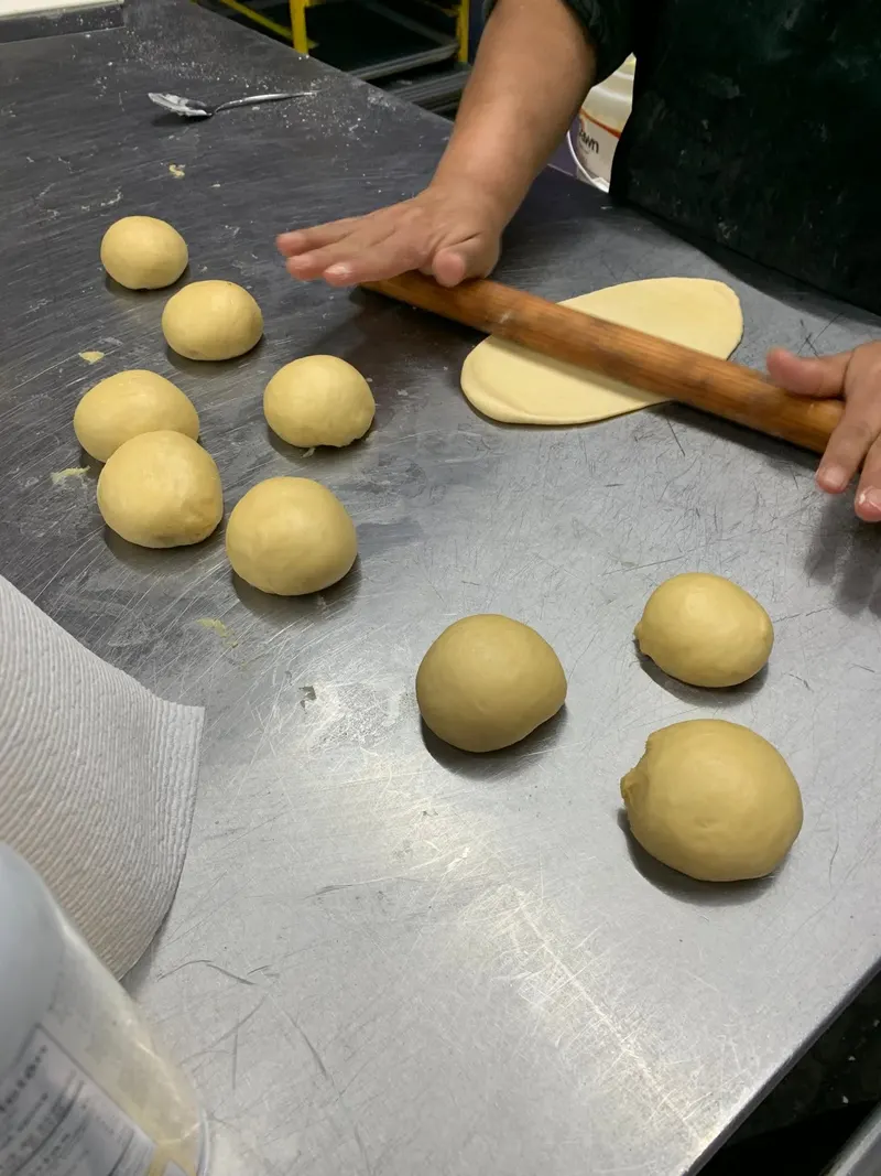 Hands rolling out bread dough on a steel table next to rounds of dough