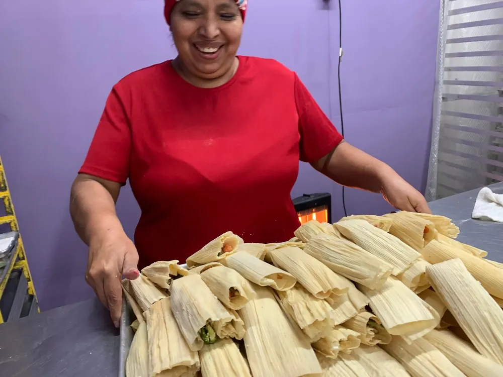 The owner of Dos Caminos Bakery smiling over a big tray of fresh tamales